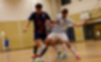 Two indoor soccer players in dark and striped jerseys battle for the ball on a gym court, with a goal visible in the background.