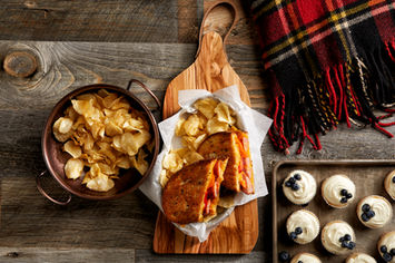 Grilled sausage on sourdough sandwich with potato chips, small cupcakes with vanilla frosting and fresh blueberries in picnic setting