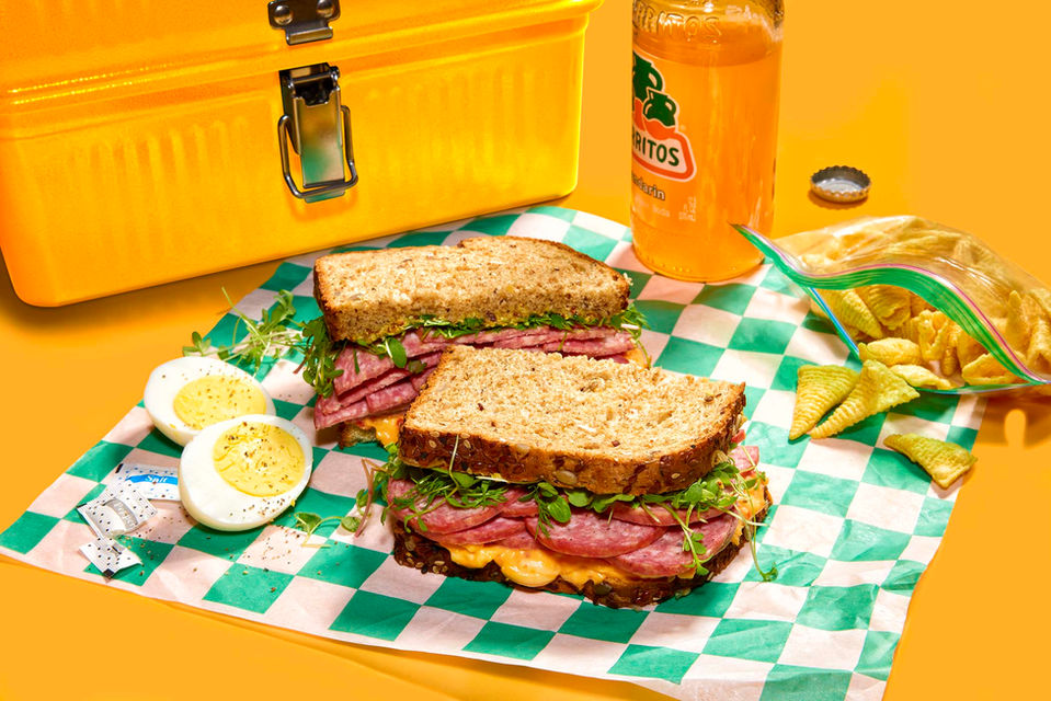 Contents of a yellow Stanley lunchbox laid out in funky retro fashion and including Johnsonville summer sausage sandwich, Bugle snacks, Jarritos orange soda and hard cooked eggs on teal checkered sandwich paper.