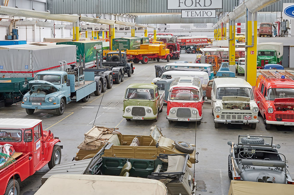 Oldtimer-LKW in einer Halle, bunte Farben. Einige Fahrzeuge mit geöffneten Motorhauben. Beschriftungen: "FORD TEMPO", "P.S. SPEICHER".