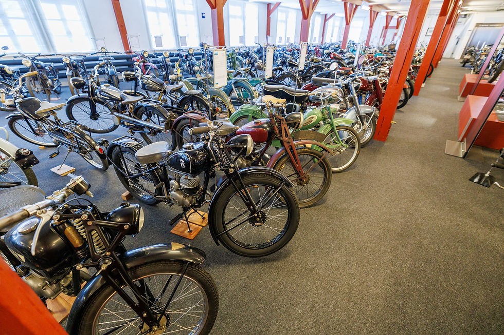Motorradmuseum voller Vintage-Motorräder in verschiedenen Farben. Holzbalken und Fenster beleuchten den Raum. Keine Menschen vorhanden.