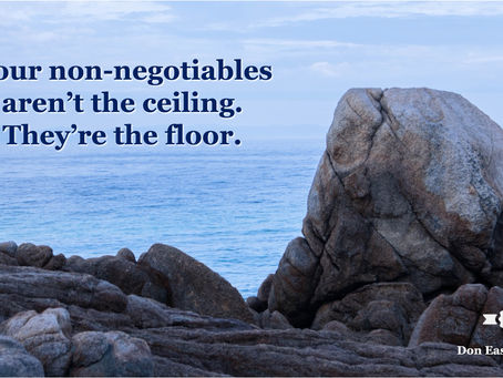 A large weathered boulder sits solidly atop a rocky coastal shoreline, with calm blue-grey ocean water stretching to a hazy horizon behind it. Bold navy text reads: "Your non-negotiables aren't the ceiling. They're the floor." Don Eash Coaching logo in the lower right corner.
