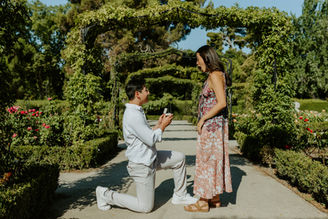 Man kneeling to propose in Retiro Park Madrid while girlfriend reacts surprised during romantic engagement photoshoot.