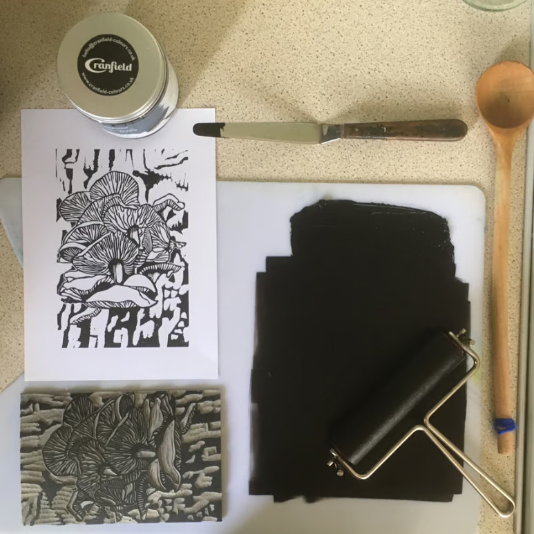 A photo of the work in progress showing the palette with ink rolled out and the roller, a palette knife, a wooden spoon, the lino and a finished print