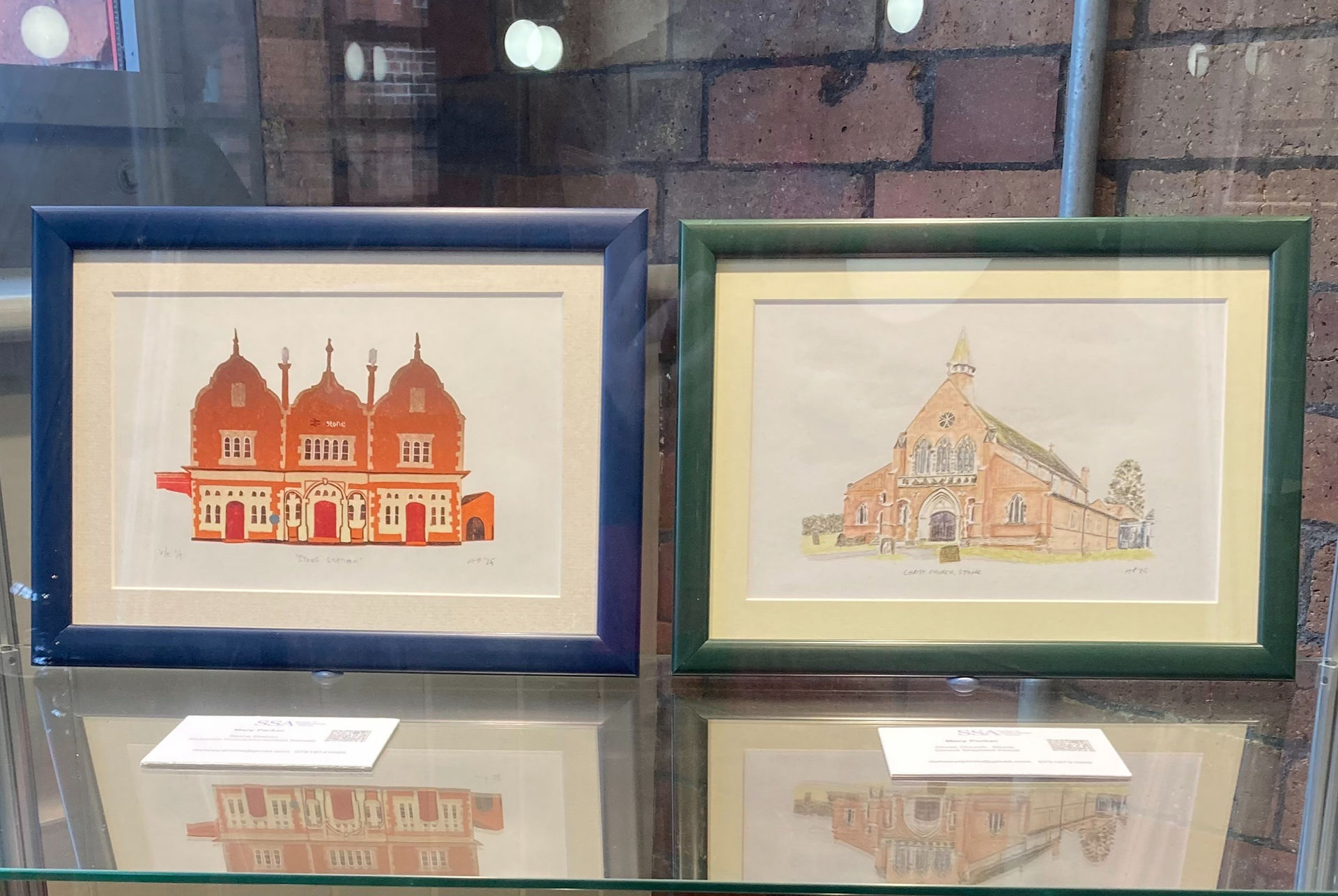 My Stone Station and Christ Church originals framed and in a glass display cabinet. Stone Station is in a navy blue frame, and Christ Church in a dark green frame. Both are reflected in the glass shelf.