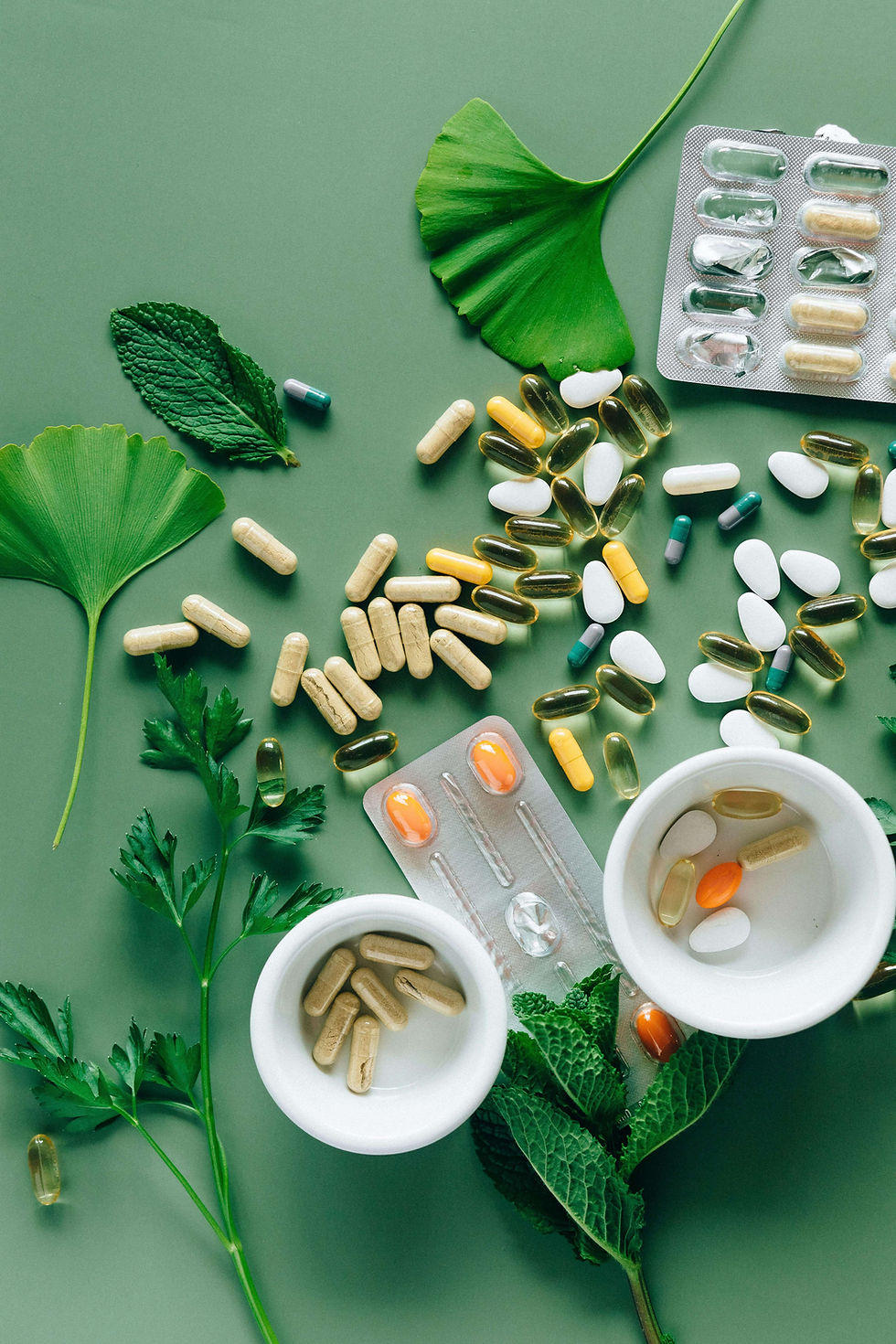 Various pills and capsules scattered on a green surface with leaves. Two white bowls hold capsules, creating a natural health theme.