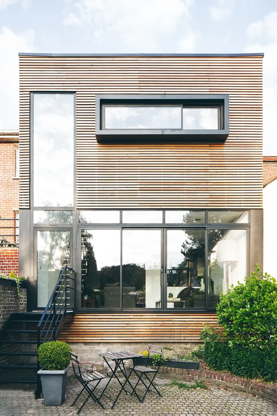 L'extension de maison émerge élégamment de son environnement, une structure cubique aux lignes épurées et contemporaines. Les larges fenêtres rectangulaires inondent l'intérieur d'une douce lumière, créant une fusion harmonieuse entre l'intérieur et l'extérieur. L'architecture audacieuse est soulignée par des matériaux modernes. Une résidence à la fois minimaliste et chaleureuse.