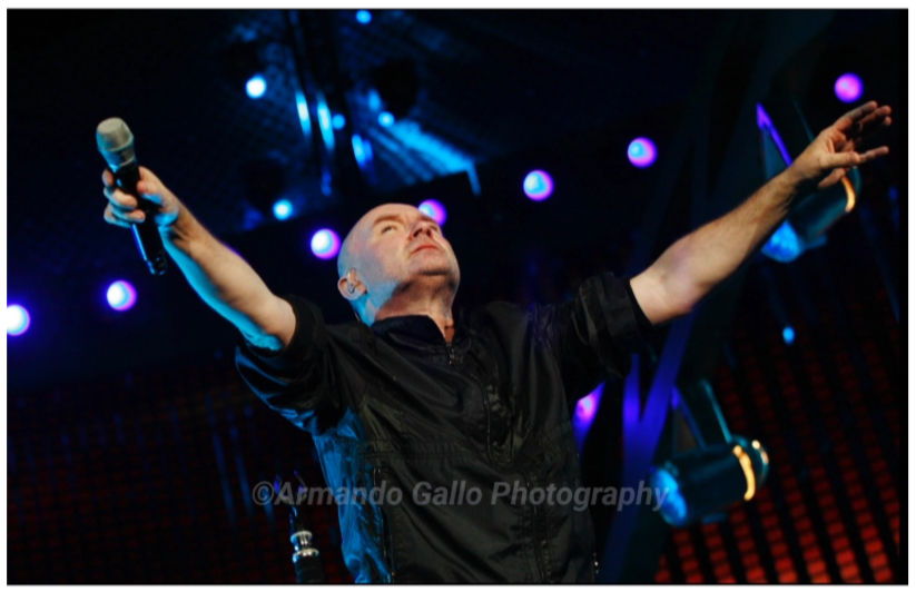 Phil Collins wearing black shirt holding microphone in his right hand arms spread out wide and up and head back with some st
