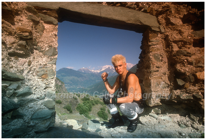 Billy Idol in sleeveless shirt crouching down on the ground on a hilltop with view through structure of the mountains