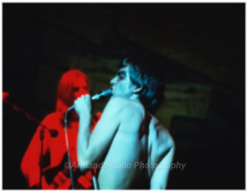 Naked back of torso blueish lighting while he holds and sings into a microphone and a guitarist in red light in background