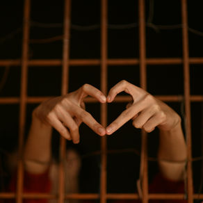 Hands form a heart shape through brown wooden bars against a dark background, conveying a sense of hope and connection.