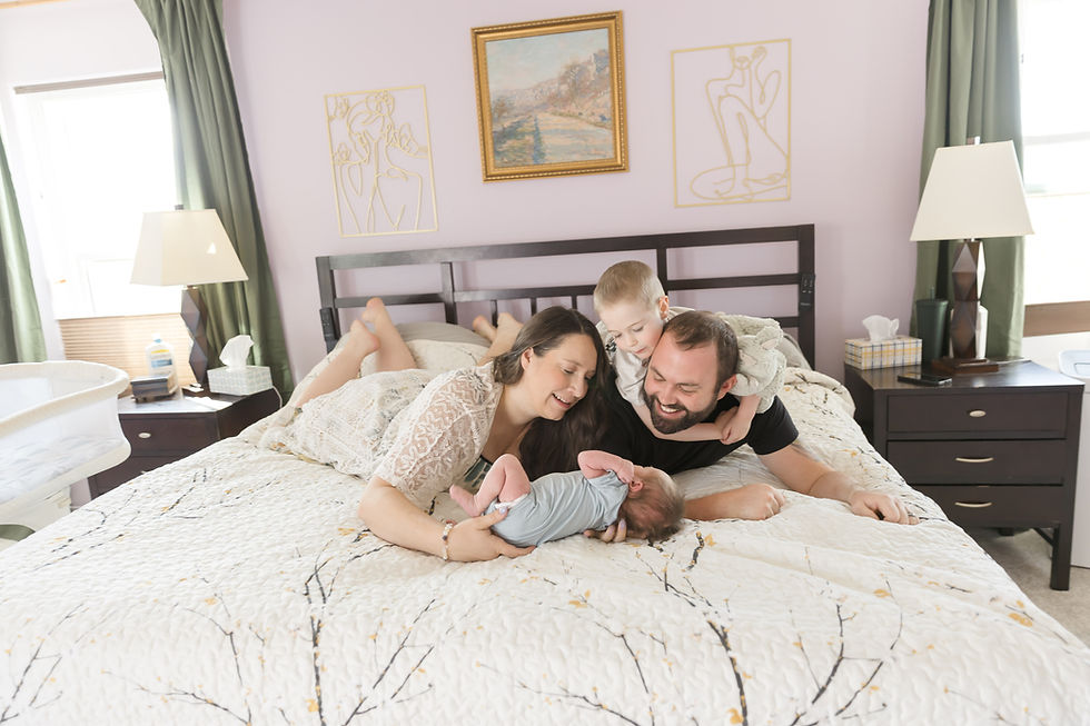Family of four on a bed; parents and young child smiling at an infant. Room with pastel walls, art, and green curtains. Cozy and joyful mood.