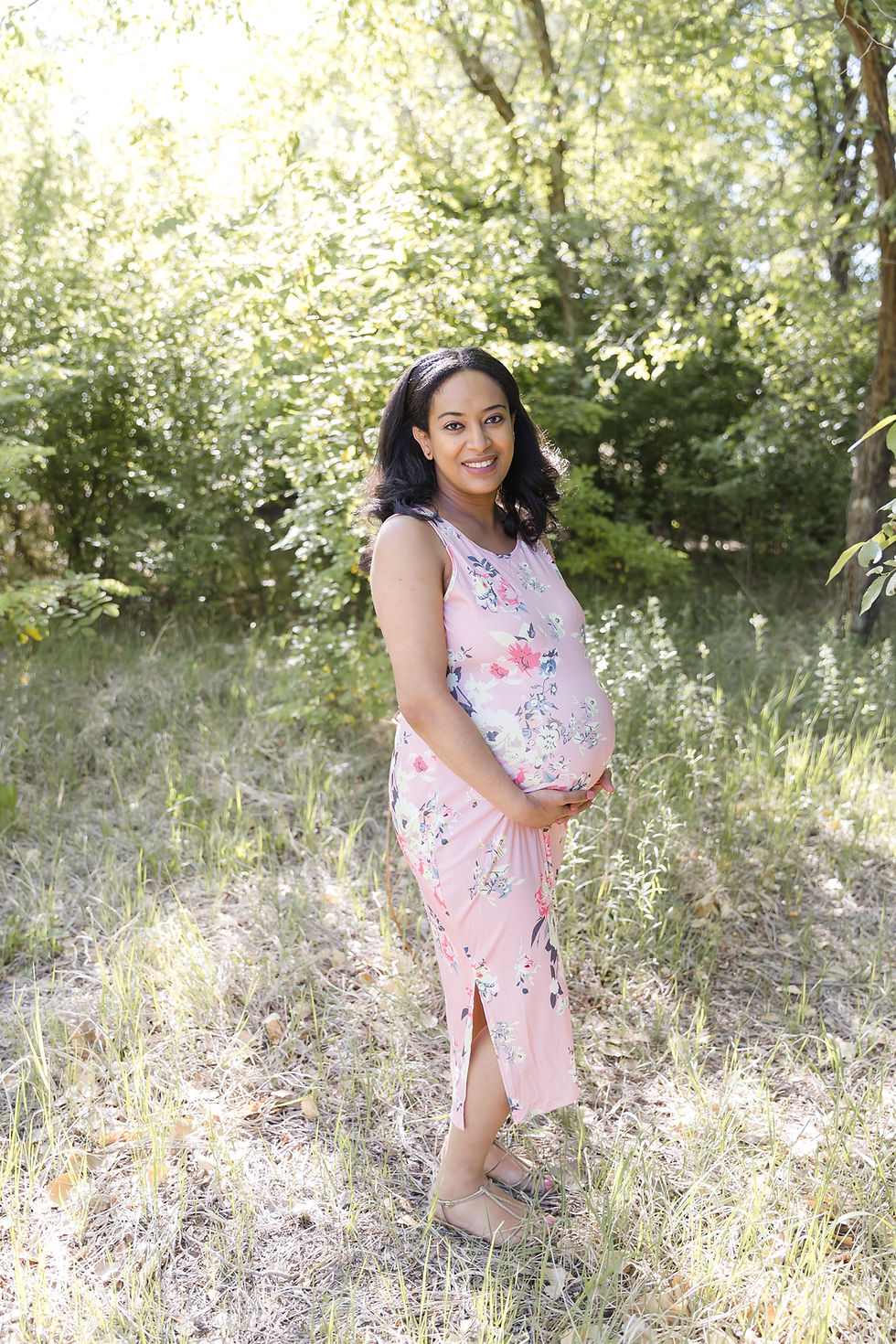 Pregnant woman in floral pink dress stands in sunny wooded area, smiling gently while cradling belly. Lush green foliage surrounds her.