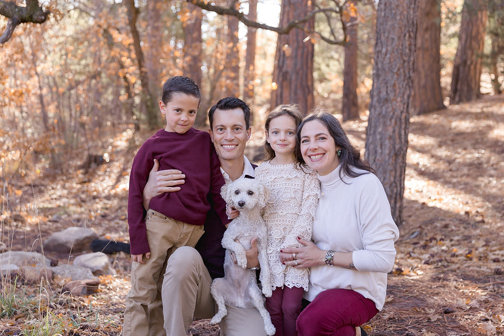 Family of four with a small white dog posing in a forest. Autumn leaves and trees surround them. Smiling faces, cozy atmosphere.