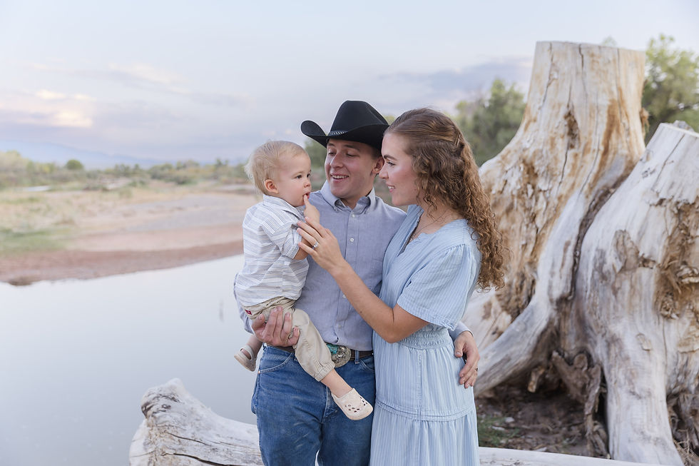 A couple with a toddler near a large driftwood trunk by a river. The man wears a cowboy hat; woman in blue dress. Warm, joyful moment.