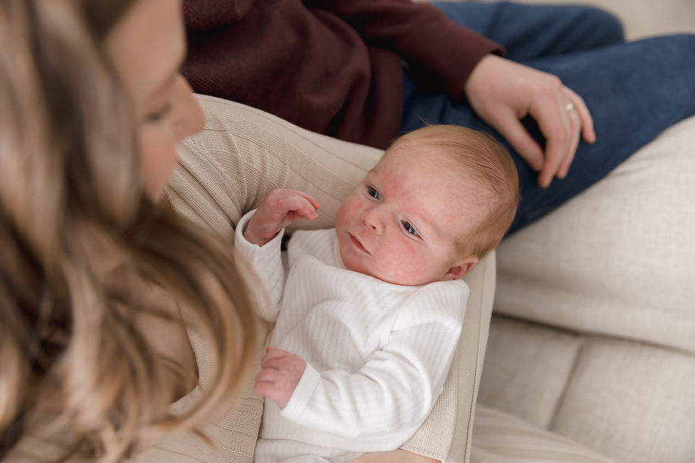 A baby in a white outfit lies in a woman's arms, while a man's hand rests nearby. Cozy, neutral-toned setting, calm mood.