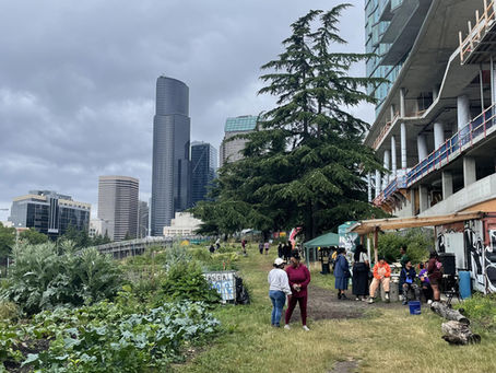 People gather near an urban garden and construction site under cloudy skies, with tall buildings in the background.