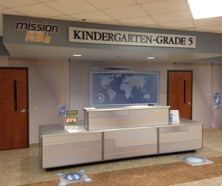 A spy themed check-in desk with a metal desk and large sign above it that reads "mission kids; kindergarten - grade five" and a map on the back wall. theme fusion