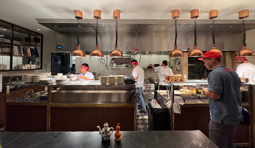 View from a seat at the chef’s table bar in the back of Norman’s restaurant, overlooking the open kitchen. Several chefs wearing white uniforms and red bandanas work under a row of copper pendant lights, plating and preparing dishes. Stacks of plates and bowls line the counter, and a chef in the foreground assembles food near the pass. The scene captures the lively, behind-the-scenes atmosphere of the kitchen from a guest’s vantage point.