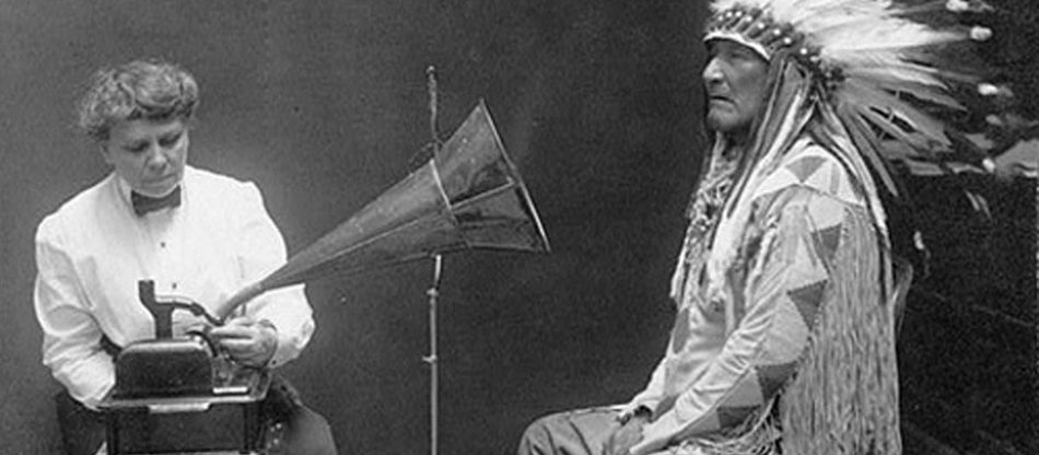 A historical black-and-white photograph of a European archivist recording an Indigenous man on a phonograph, used here as a metaphor to critique 'Native-Speakerism'—the archaic and discriminatory bias that prioritizes a teacher's birthplace over their professional expertise and pedagogical skill.