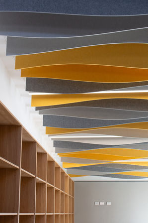 Curved suspended acoustic ceiling fins in alternating grey, white and yellow colours beneath a perforated ceiling.