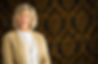A smiling real estate agent in a beige cardigan and white shirt stands against an elegant black and gold patterned background.