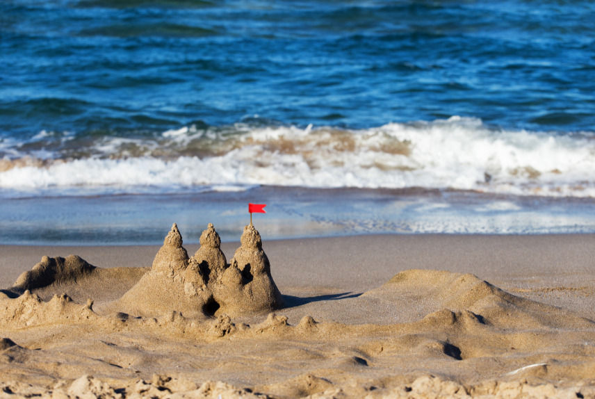 A sand castle with a red flag being washed away slowly by the sea.