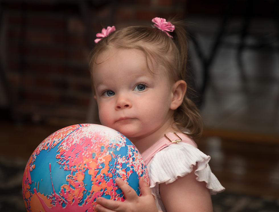 Toddler with pink bows holds a colorful ball in a cozy room with a brick wall. She looks curious, wearing a white ruffled dress.