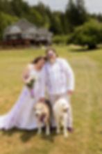 A woman in a flowing white and lavender wedding dress holds a bouquet, smiling outdoors near a forest and lake. Her head is on her husband's shoulder A cozy house is nearby. Seabeck Wedding