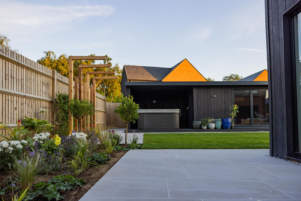 A porcelain patio and natural grass area by a flowerbed and a garden room with a jacuzzi at the end