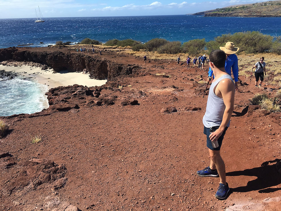 Man in tank top and shorts stands on rocky cliff, looking at hikers approaching. Ocean and sailboat visible in the sunny background.