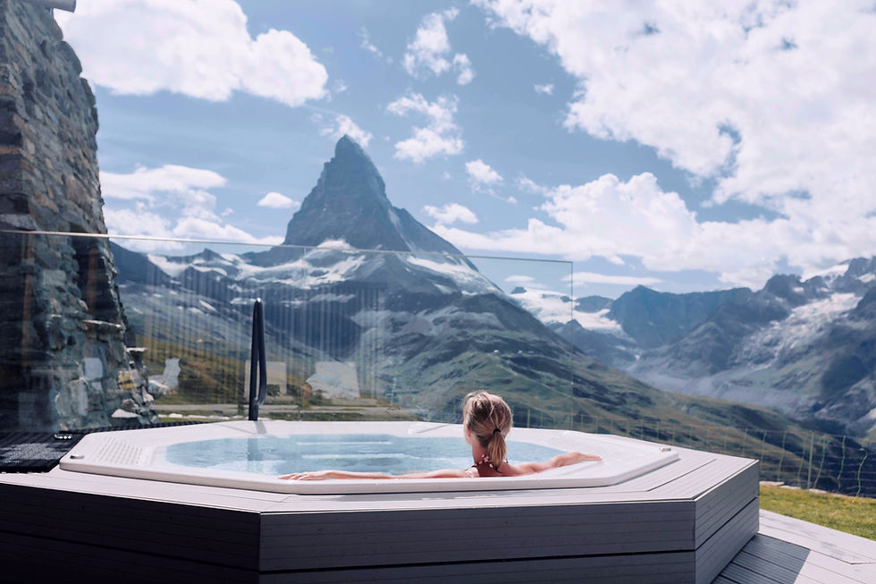 Person relaxing in a hot tub with arms outstretched, overlooking the Matterhorn. Clear blue sky with scattered clouds. Serene and calm mood.