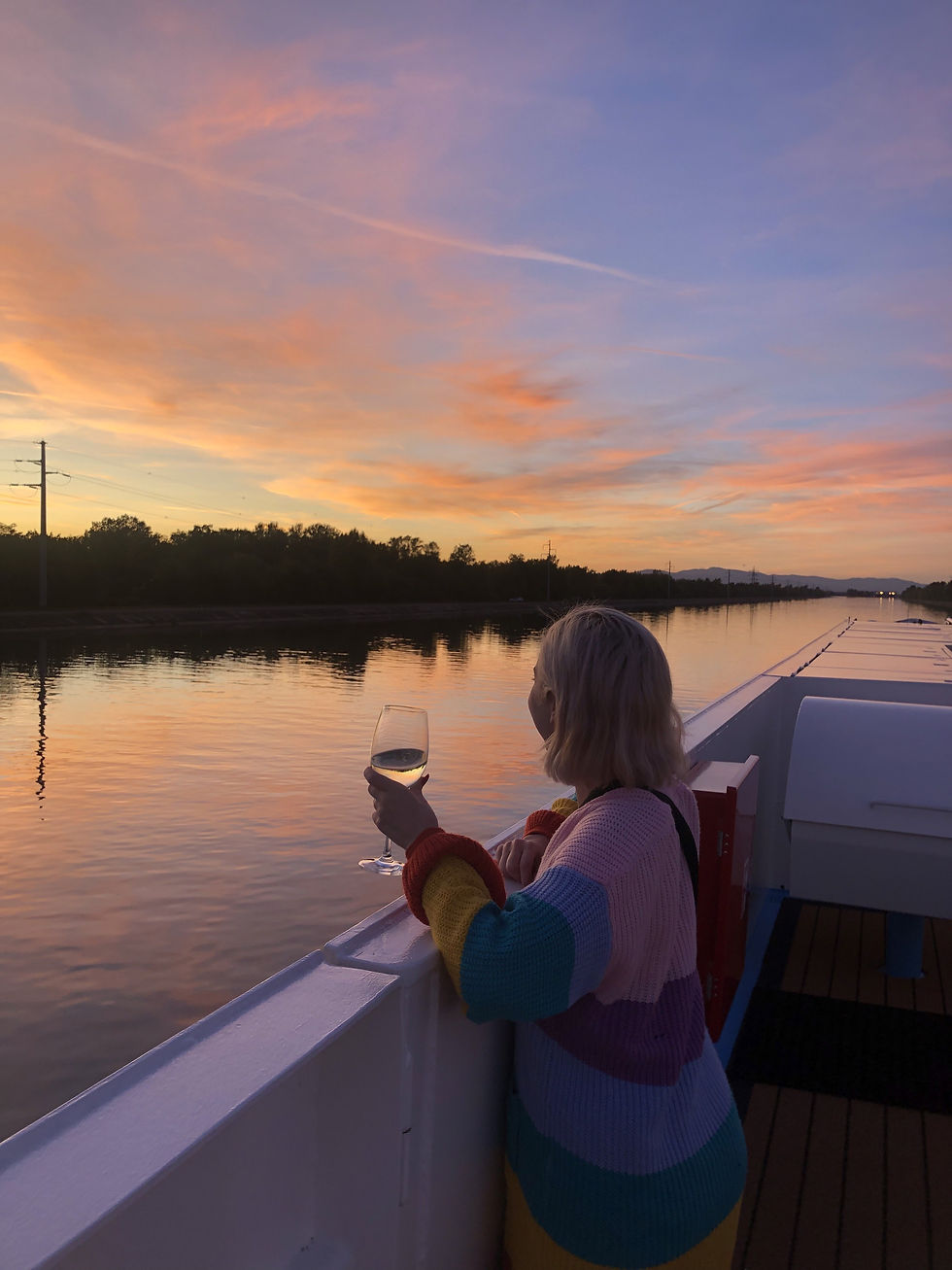 Woman in a rainbow sweater holds a wine glass on a boat, gazing over calm water at a pastel sunset.