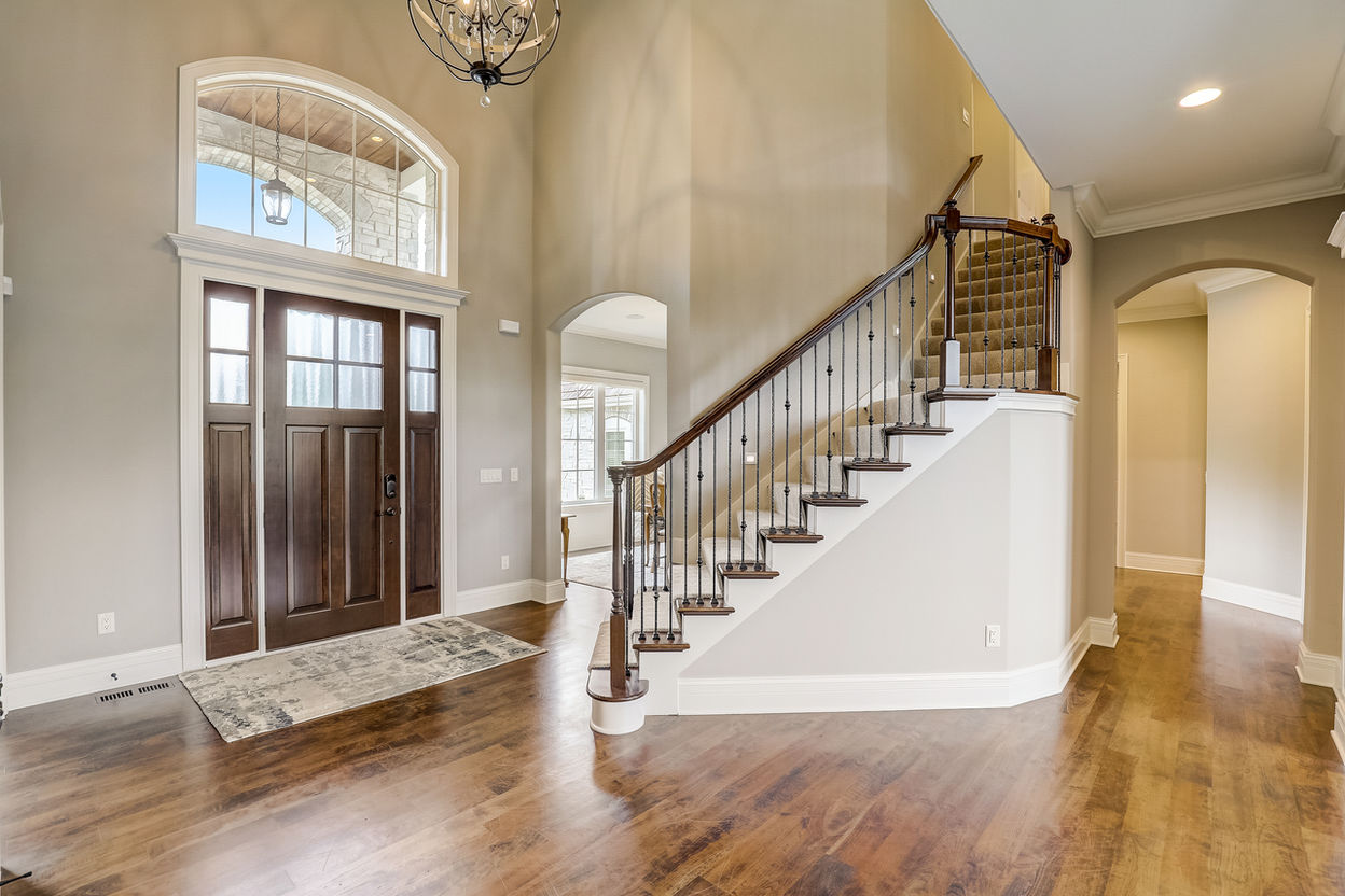 Tall wooden front door with large window above and sidelights, staircase up to second floor, custom hardwood flooring.