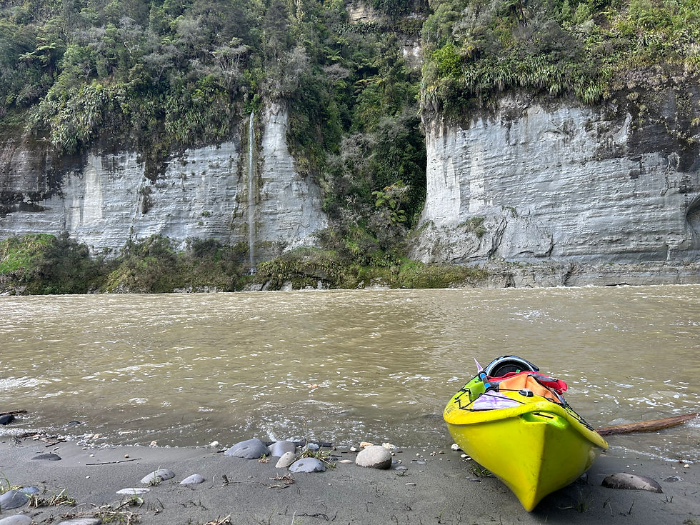 A yellow kayak sits on a riverbank facing a rocky cliff topped with greenery.