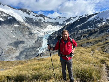 Author Will Duncan stands on a grassy hill in front of a large rocky glacier on a bright and sunny day.