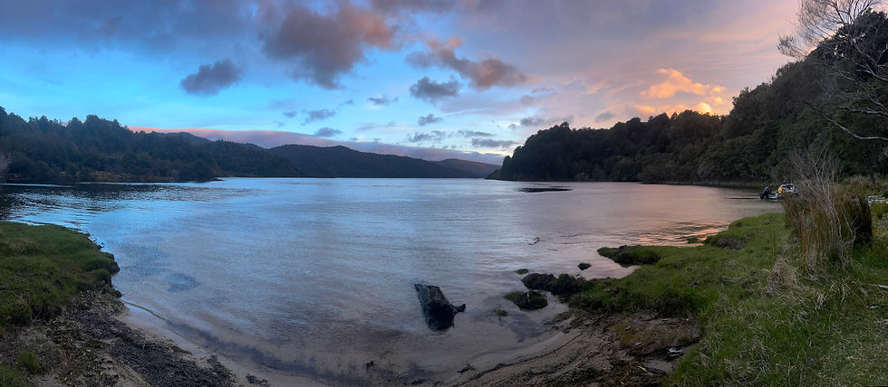 A panoramic view of a lake as the sun is setting. The sky is blue to the left and orange to the right.
