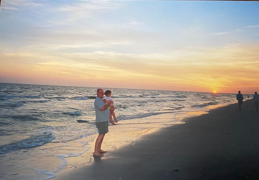 A young Rebekah carried by her father who is wearing a light blue shirt and darker shorts. They are standing on a beach as the waves roll in and the sun is setting in the background.