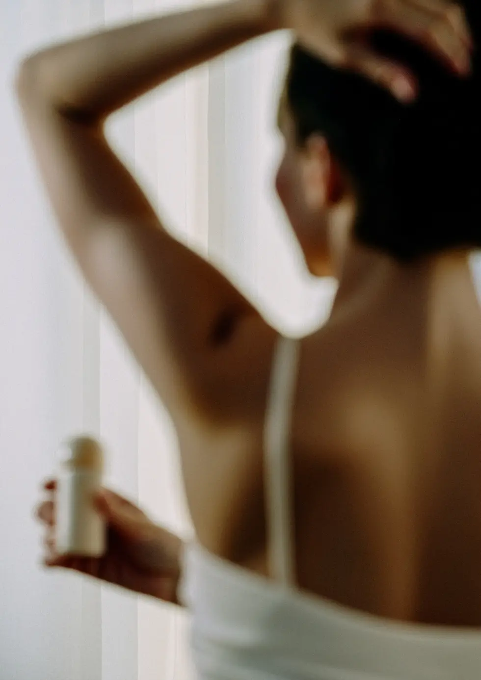 Thumbnail: The back of a woman applying Pure Deo Natural Refillable Roll On Deodorant by a window with curtain pulled