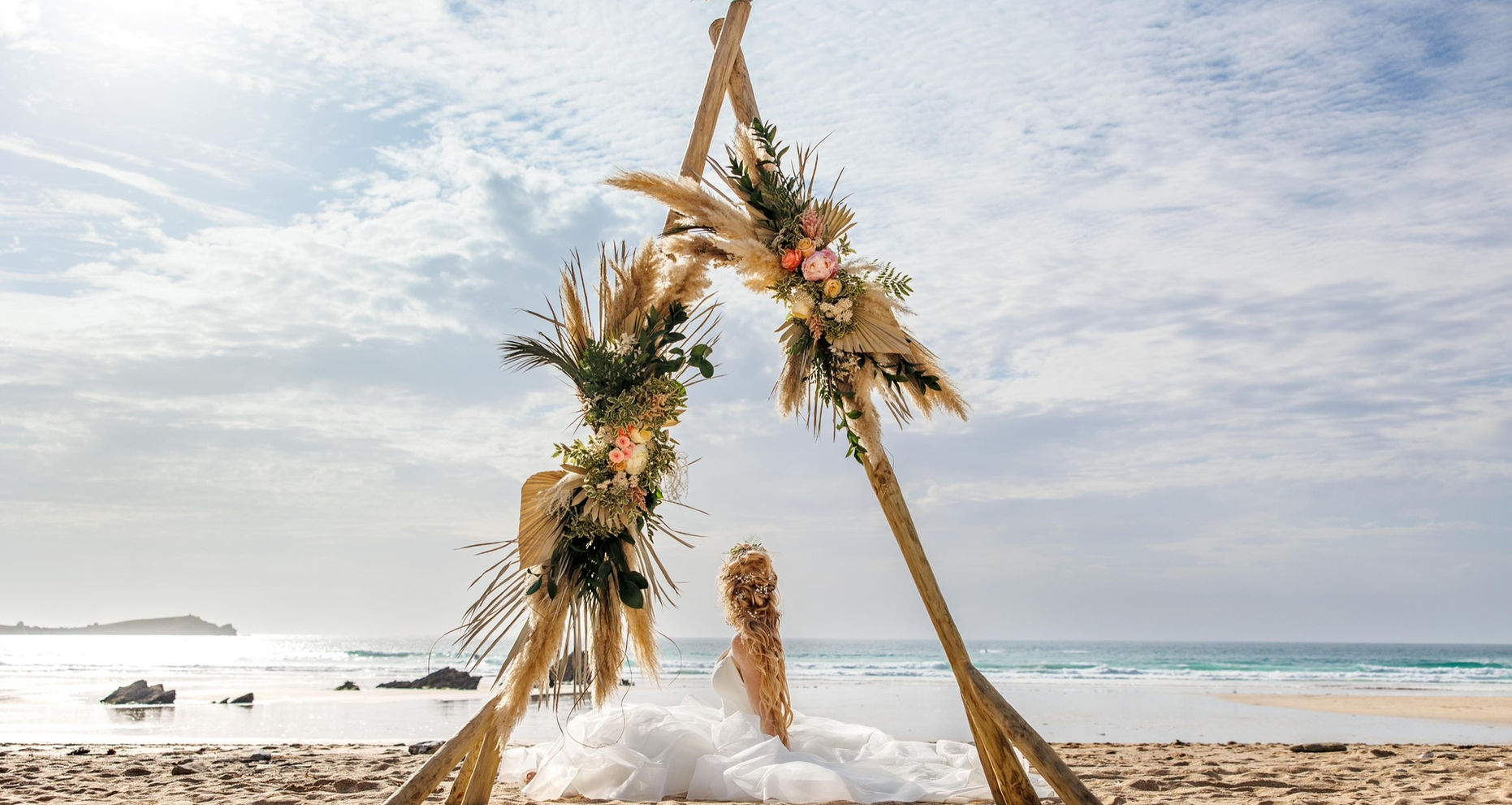 A bride seated on a beach under a triangular driftwood arch adorned with pampas grass and flowers, created by award-winning florist Blossom and Kelp.