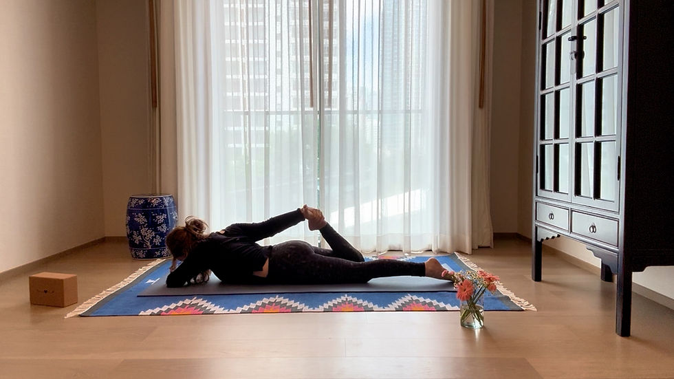 Person in black outfit doing yoga on a colorful mat indoors, near a cabinet and flowers. Bright window in background. Peaceful mood.