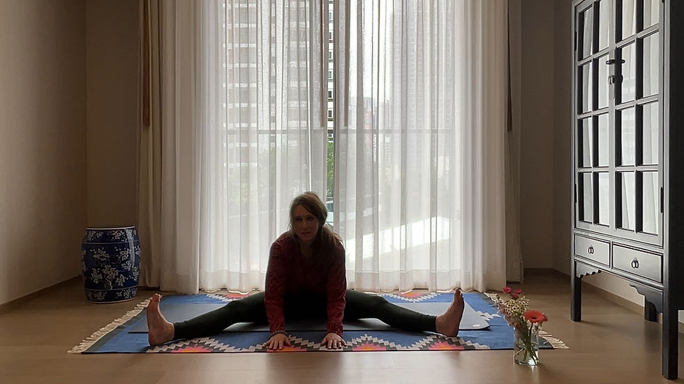 Woman in yoga pose on a colorful mat indoors, by a window with sheer curtains. Room has a blue vase, flowers, and a cabinet.