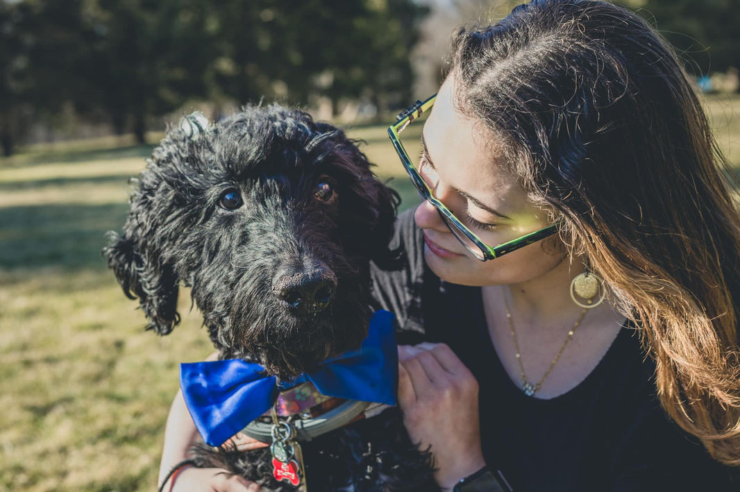 A young white woman with dark brown hair, wearing glasses, gold hoop earrings, and a black shirt, wraps her arms around her black Labradoodle, which is wearing a royal blue bow tie and guide harness.