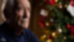 Elderly man gazes pensively beside a decorated Christmas tree with red baubles and stockings, set in a warm, festive ambiance.