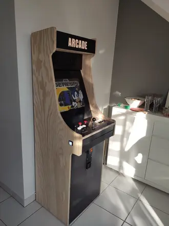 Machine d’arcade bois brut à deux joueurs, dans un décor minimaliste avec plante verte et tapis en jute, jeux rétro inclus