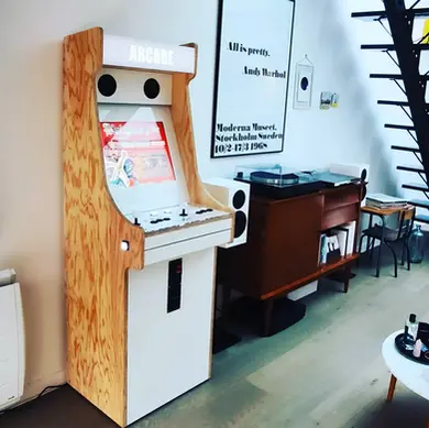 Borne d’arcade en bois brut deux joueurs, installée dans un salon design avec jeux rétro comme Pac-Man et Donkey Kong