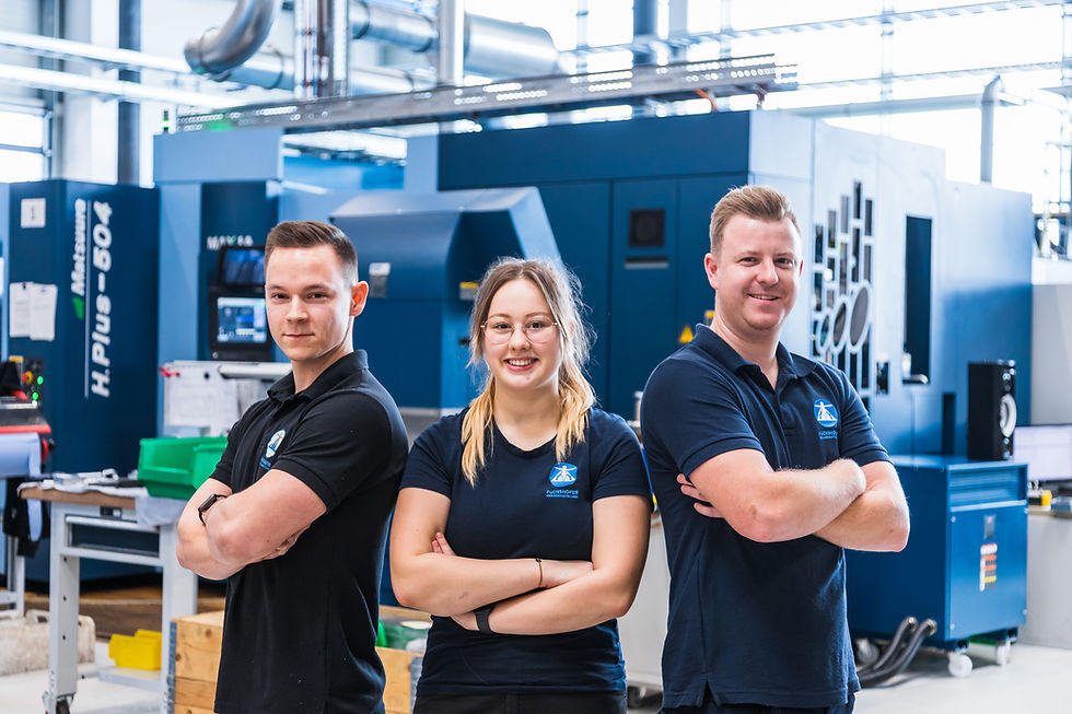 Drei Mitarbeitende von Fuchshofer Präzisionstechnik stehen selbstbewusst mit verschränkten Armen in einer modernen Werkhalle. Links ein junger Mann, in der Mitte eine junge Frau mit Brille, rechts ein Mann mit hellem Haar. Alle tragen dunkle Firmenpoloshirts mit Logo. Hinter ihnen stehen große blaue CNC-Maschinen und Werkstattausrüstung.