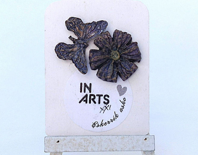 Par de brincos assimétricos em pasta de papel com uma borboleta (2,5 cm) e uma flor (2 cm) em tons de roxo e dourado, expostos num cavalete branco.