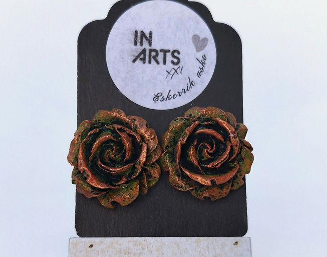 Par de brincos em forma de rosa em pasta de papel com acabamento cor cobre e detalhes de pátina verde, 2,5cm de diâmetro, sobre cartão preto.