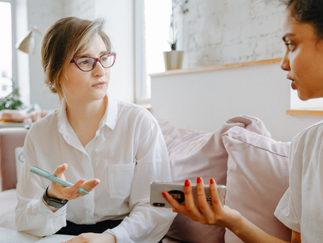 Two women talk on a couch in a bright living room, one holding a pen and notebook, the other a phone.